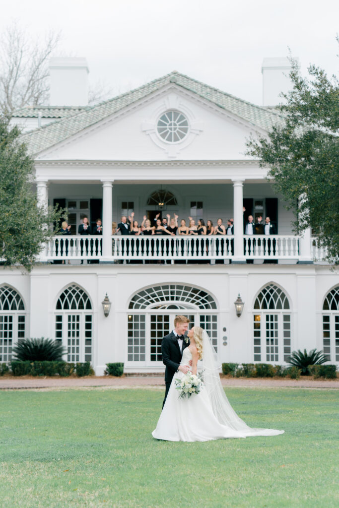 Bride and groom looking at each other in front of Lowndes Grove with bridal party cheering from the porch. 