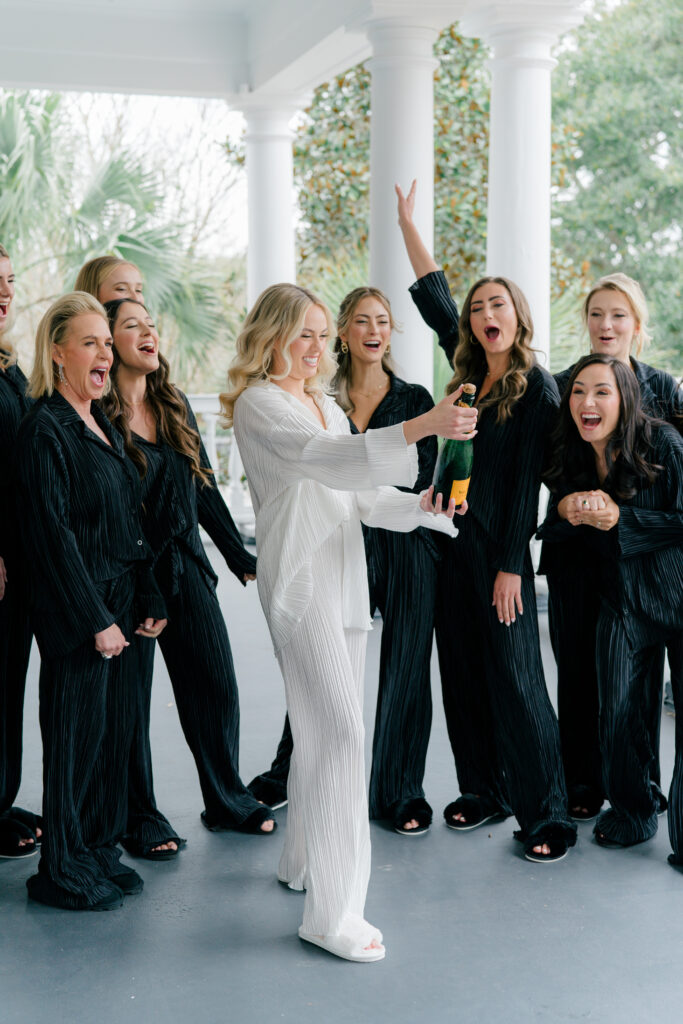 Bride popping champagne on the porch with bridesmaids in black pajamas cheering.