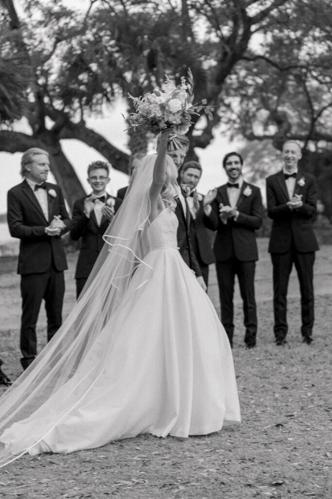 Black and white photo of bride raising bouquet with groomsmen clapping in the background. 