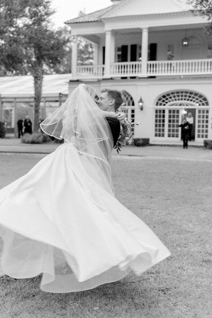 Black and white photo of groom picking up bride and spinning her on the front lawn of Lowndes Grove. 