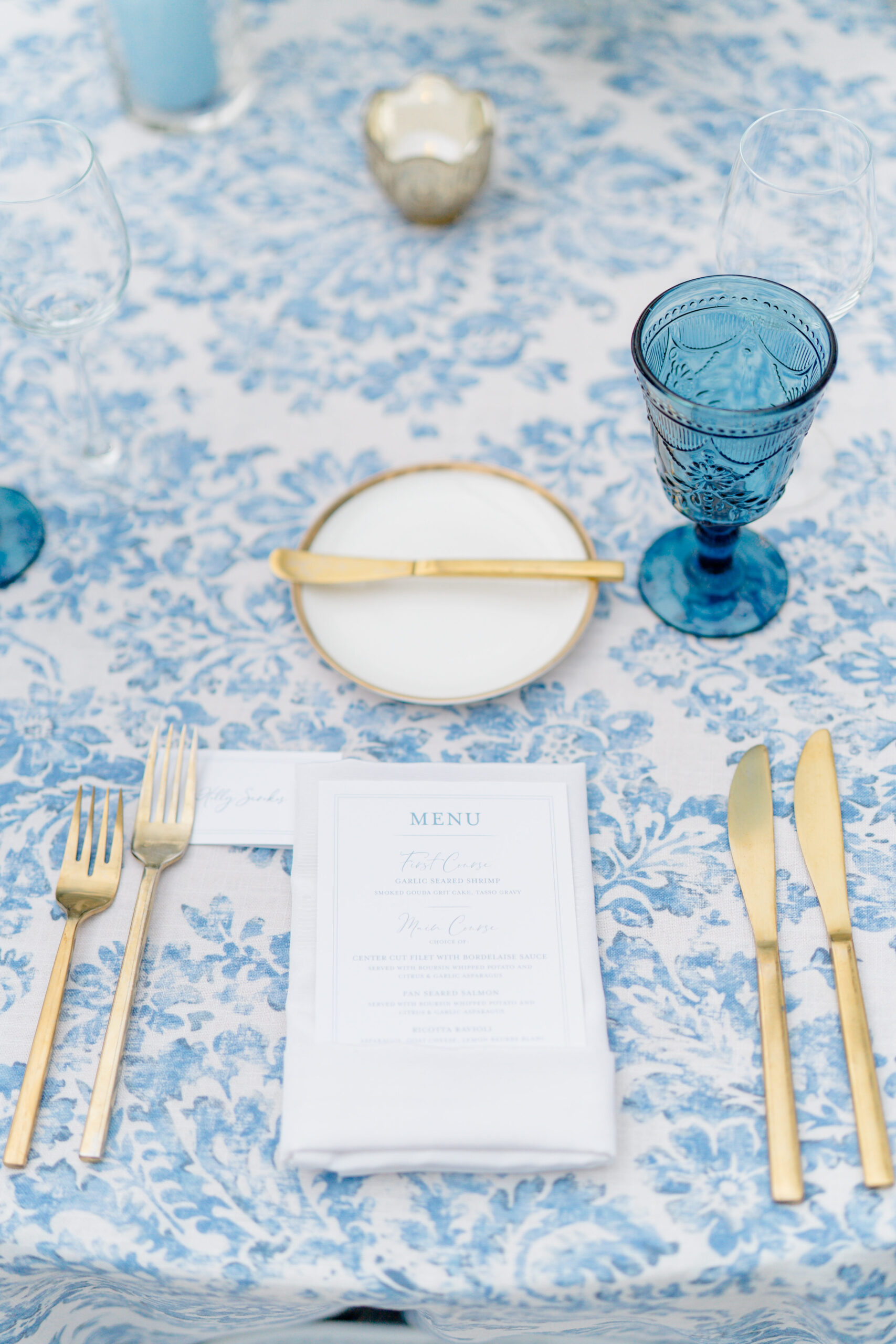 Blue and white floral table cloth with gold utensils and blue glass chalice.