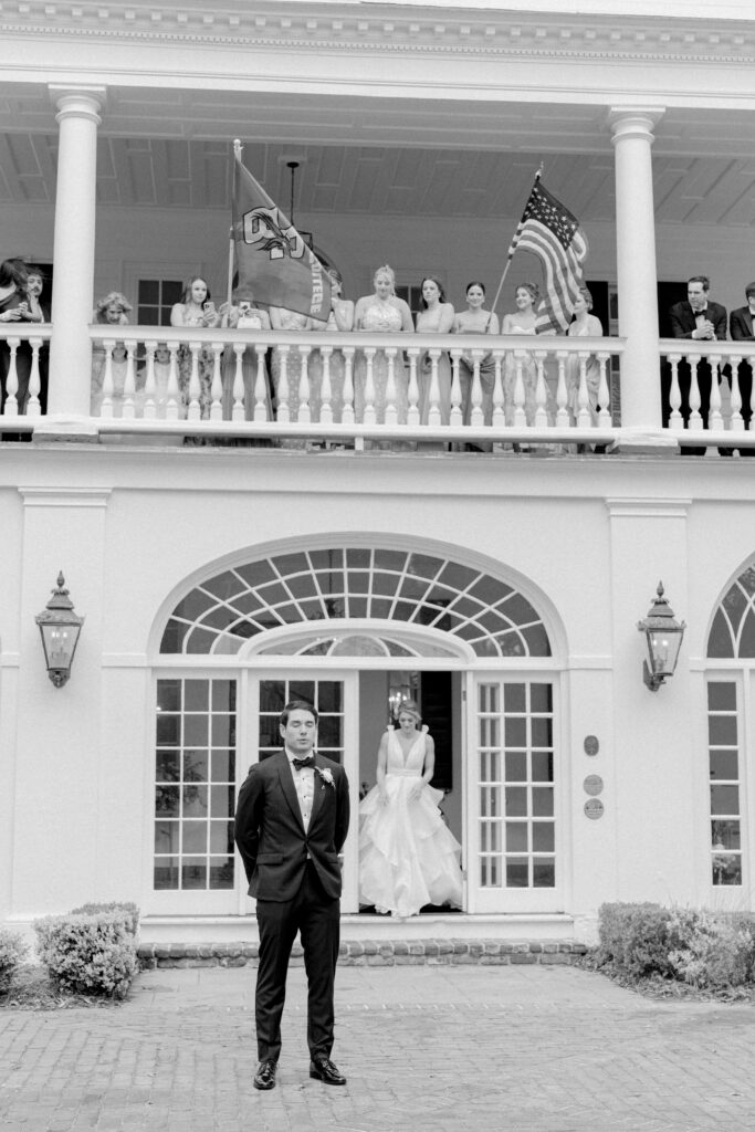 Bride walks out the door at Lowndes Grove with groom waiting for her. First look. Bridesmaids watching from the porch. Black and white wedding photo.