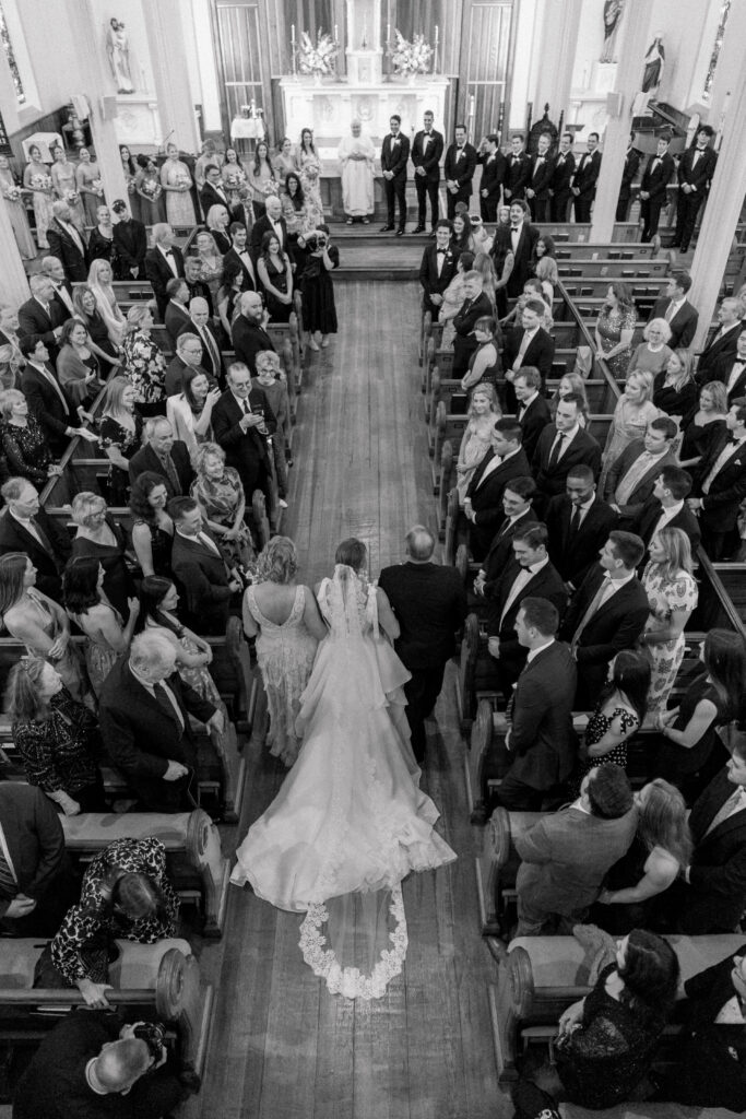 Black and white photo of above. Bride walked up the aisle by her parents with all wedding guests turned around to see her coming up the aisle. 