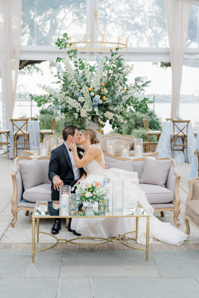 Big statement pedestal flowers with bride and groom kissing on a couch in front of it. Wedding reception. Charleston photographer.