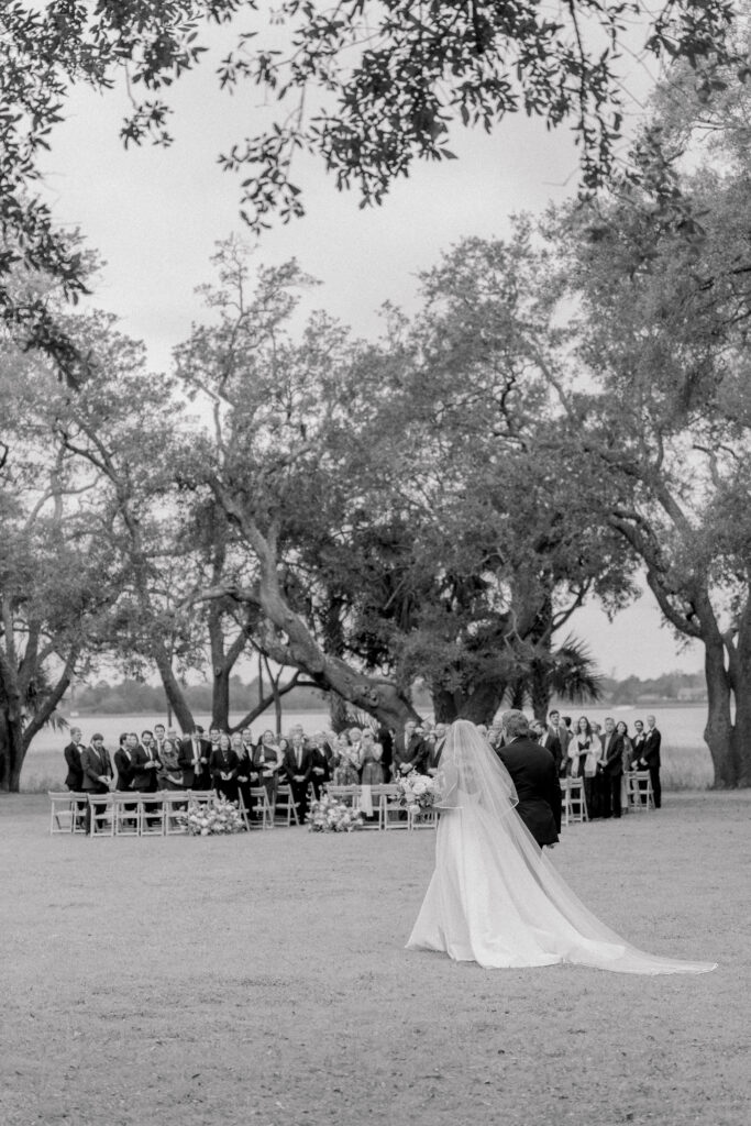 Black and white photo of bride and dad walking to wedding ceremony with guests in the background. 