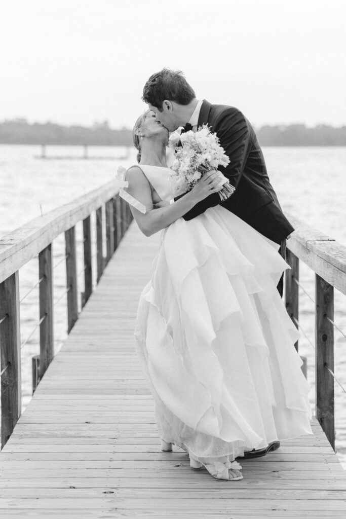 Black and white photo of bride and groom dip kiss on the dock at Lowndes Grove spring wedding. 