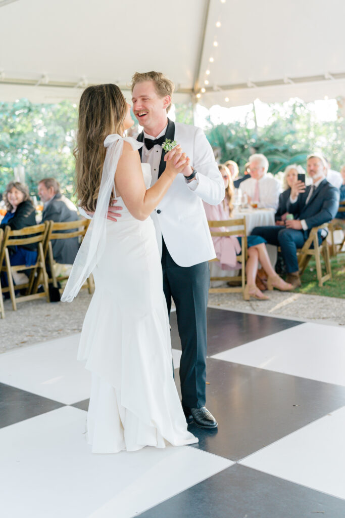Groom laughs with bride during first dance on checkerboard dance floor.