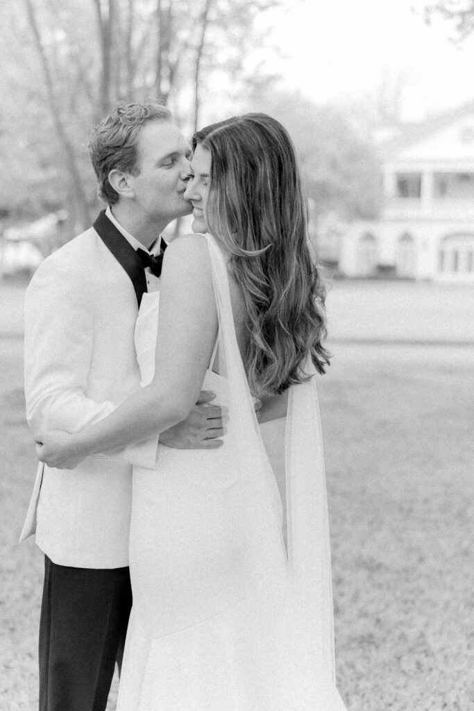 Groom in white tuxedo jacket kisses bride's cheek on the lawn in front of Lowndes Grove. Black and white photo.