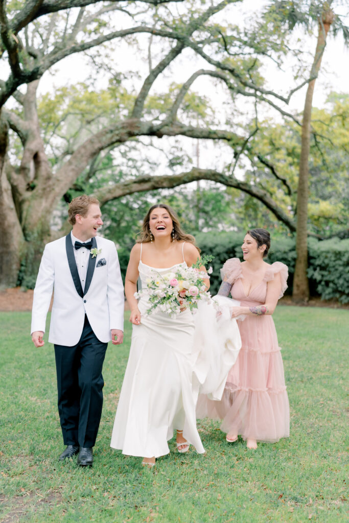 Maid of honor helps bride with her dress. Groom in white tuxedo jacket walking with bride and maid of honor. 