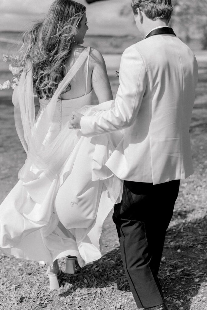 Black and white photo of groom helping bride with wedding dress while walking.