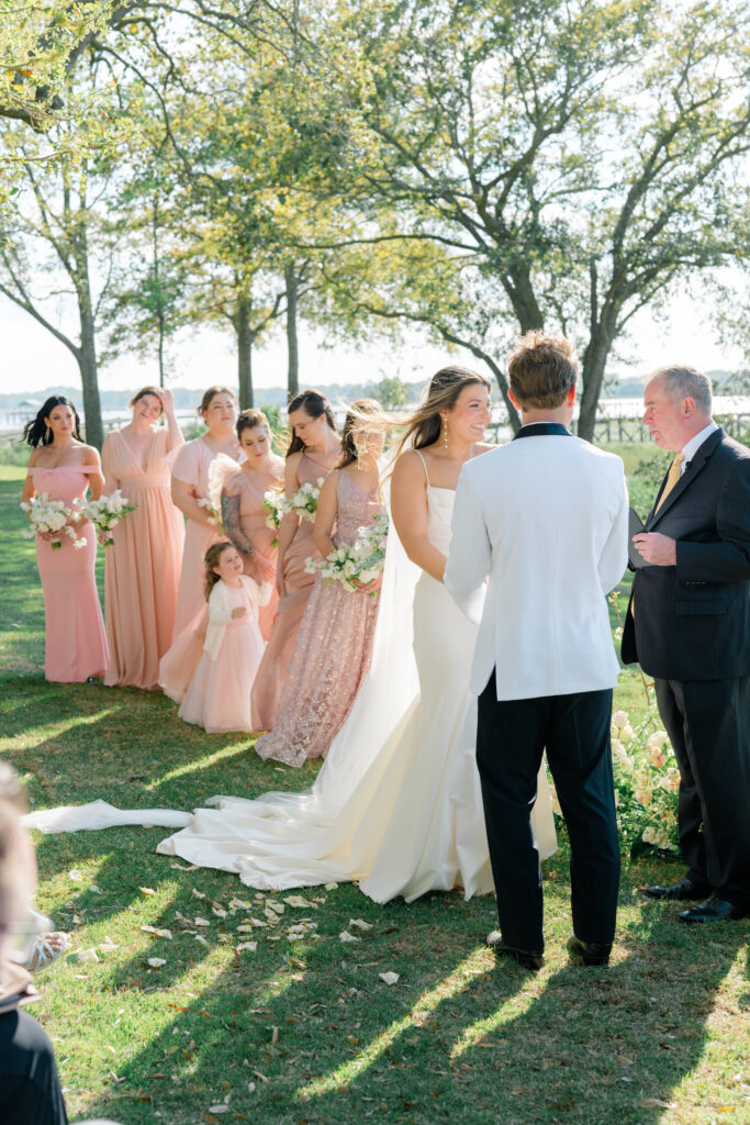 Bride and groom standing at wedding ceremony with bridesmaids in pink dresses in the background. River House at Lowndes Grove. 