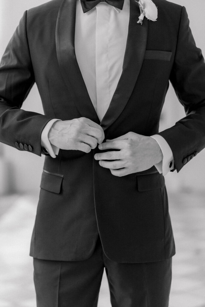 Black and white photo of groom's hands buttoning his tuxedo jacket. 