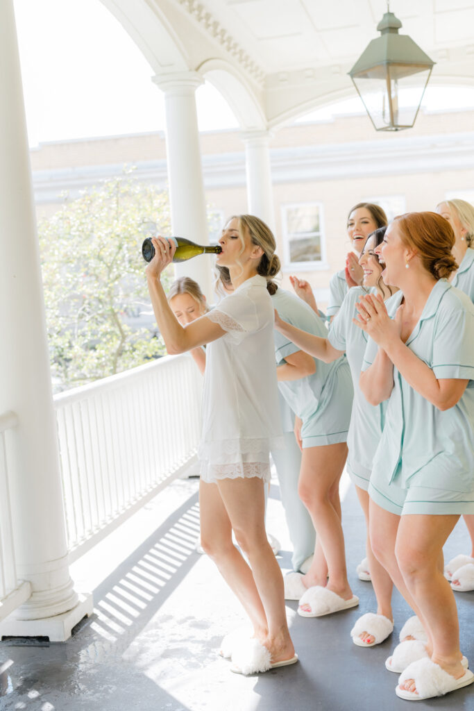 Bride sips champagne right out of the bottle on the porch with bridesmaids.