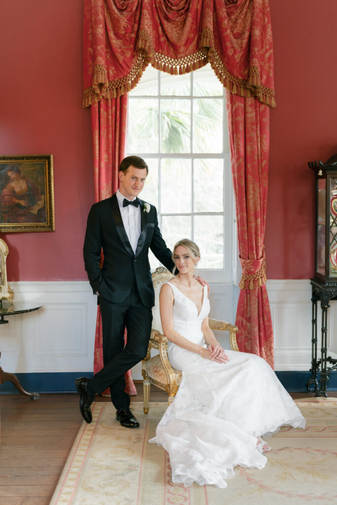 Bride sitting and groom standing. Portraits in the red room at William Aiken House. Historic Charleston wedding. 