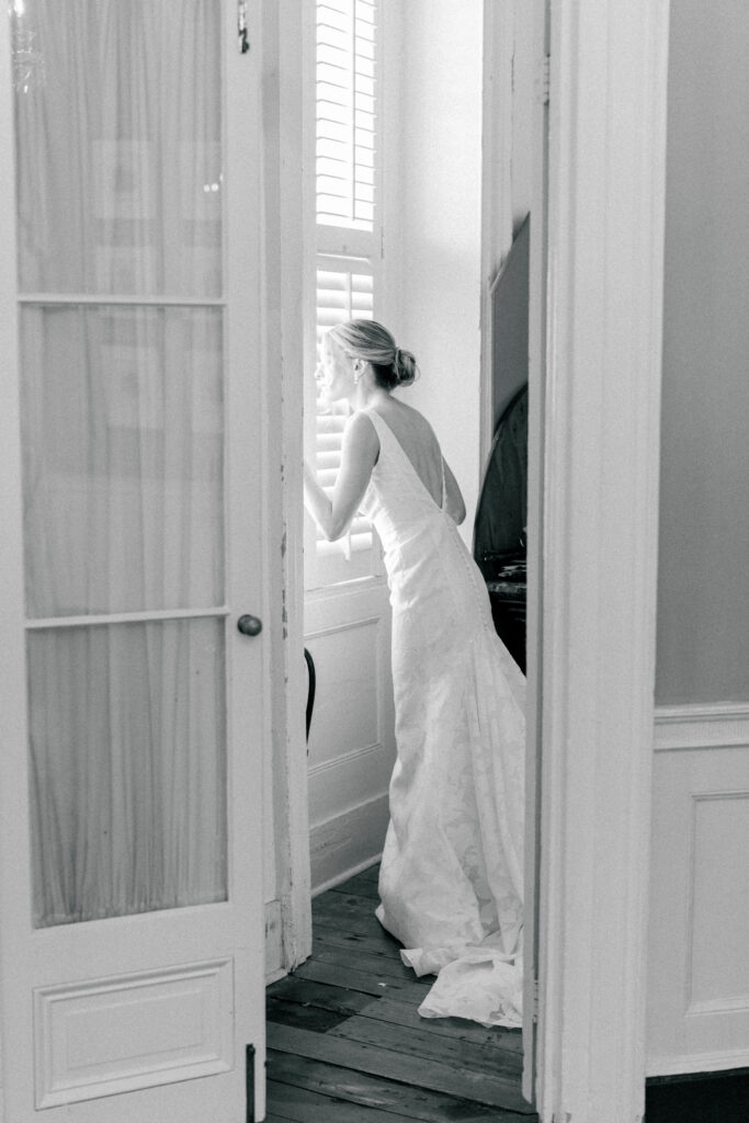 Black and whit photo of bride in wedding dress peeking out the window.
