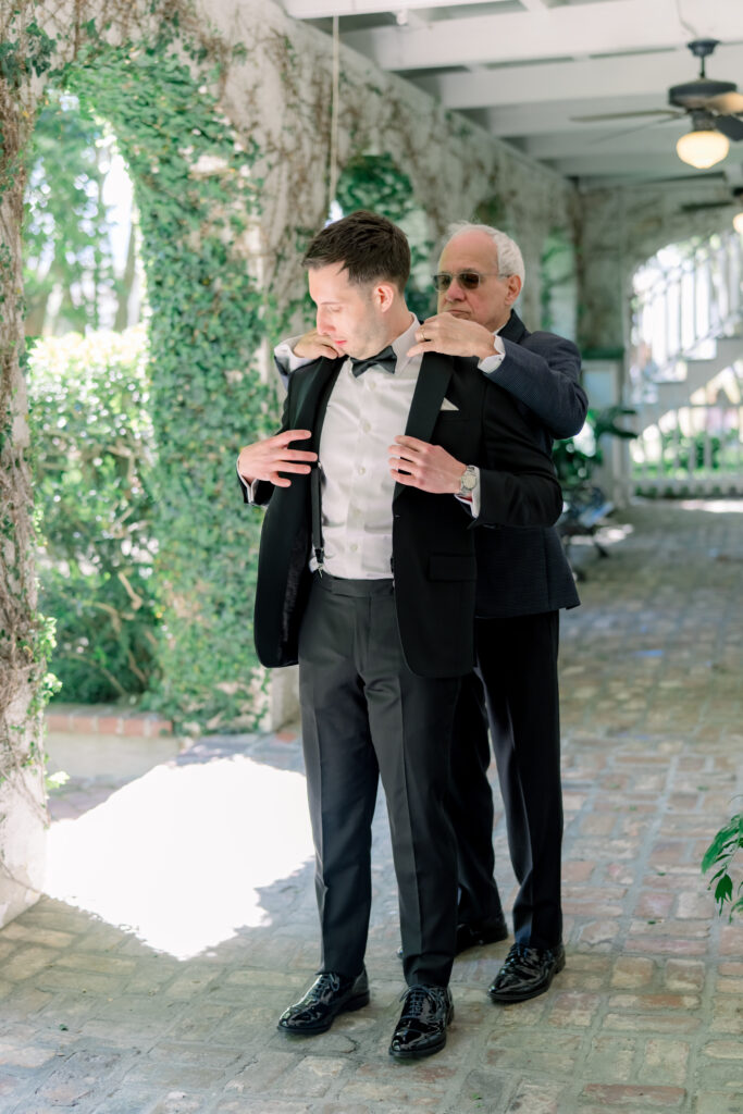 Groom puts on his tuxedo jacket with help from his dad. 