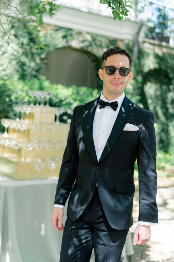 Candid photo of groom walking during wedding day with champagne tower in background. 