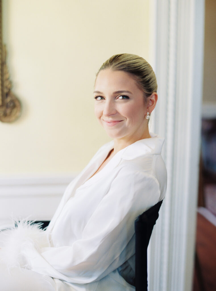 Bride in makeup chair on the morning on her wedding with white pajamas and fluffy cuffs. 