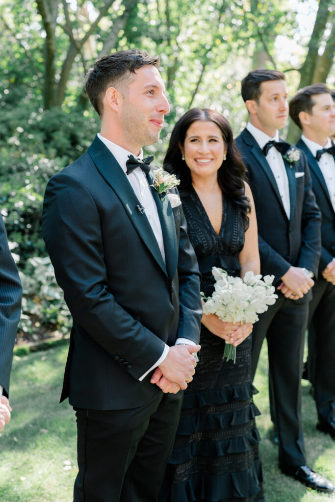 Emotional groom seeing the bride at outdoor wedding ceremony with his sister smiling and looking at him. 