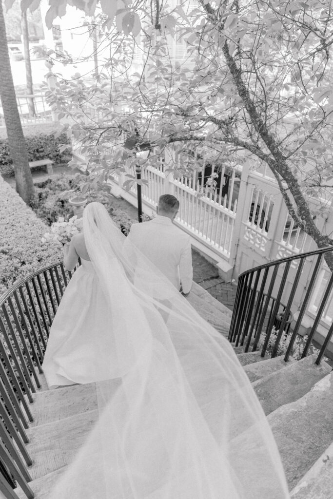 Black and white photo of bride being escorted by her dad down curved marble steps with veil blowing in the wind. 