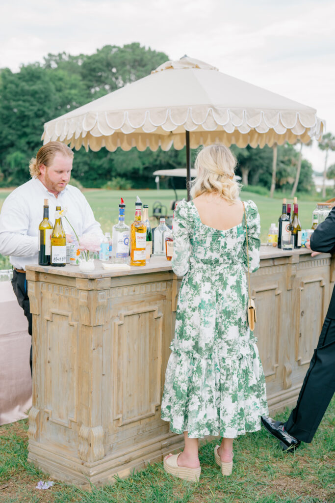 Wedding guest in green floral dress at wooden bar with scalloped tan umbrella. 