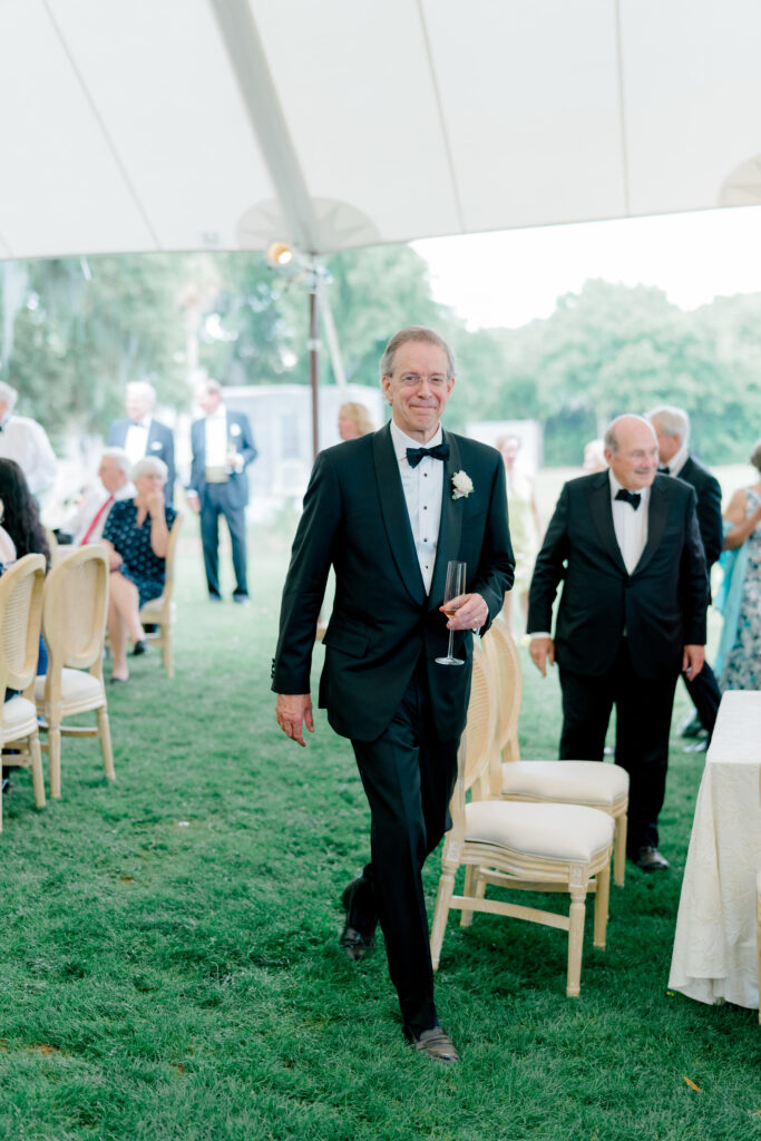 Father of the groom entering wedding reception tent holding glass of champagne. 