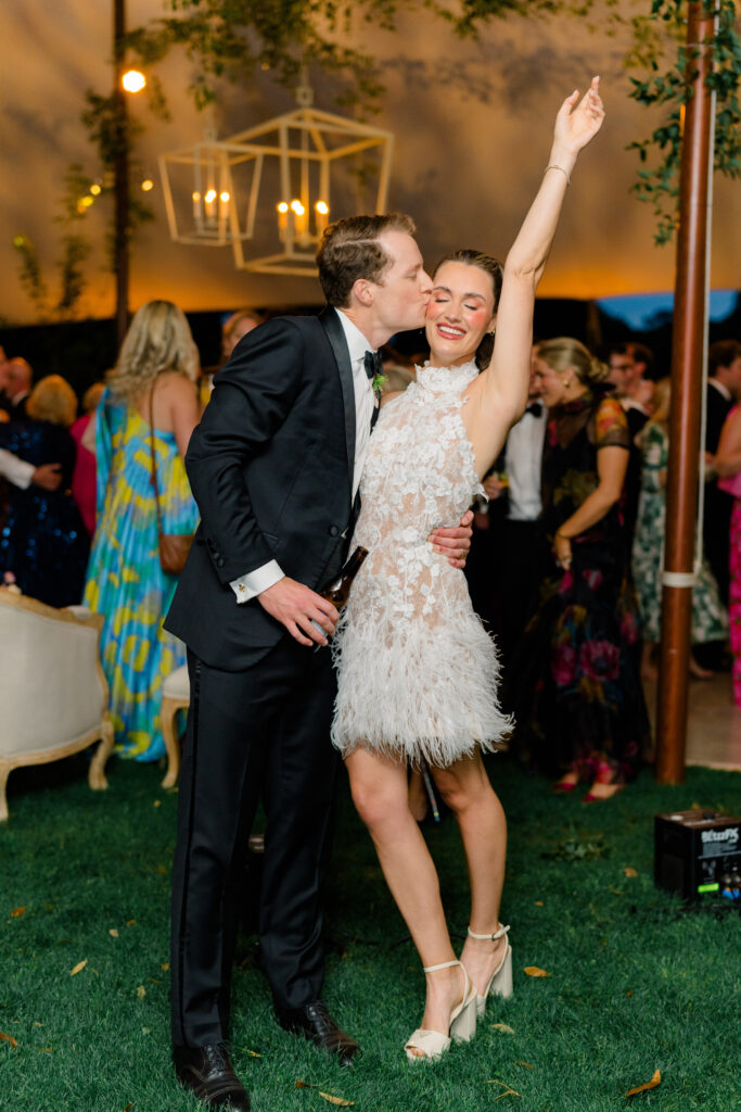 Bride changes into high neck second dress. Groom kisses brides cheek and bride smiles with her arm in the air. 