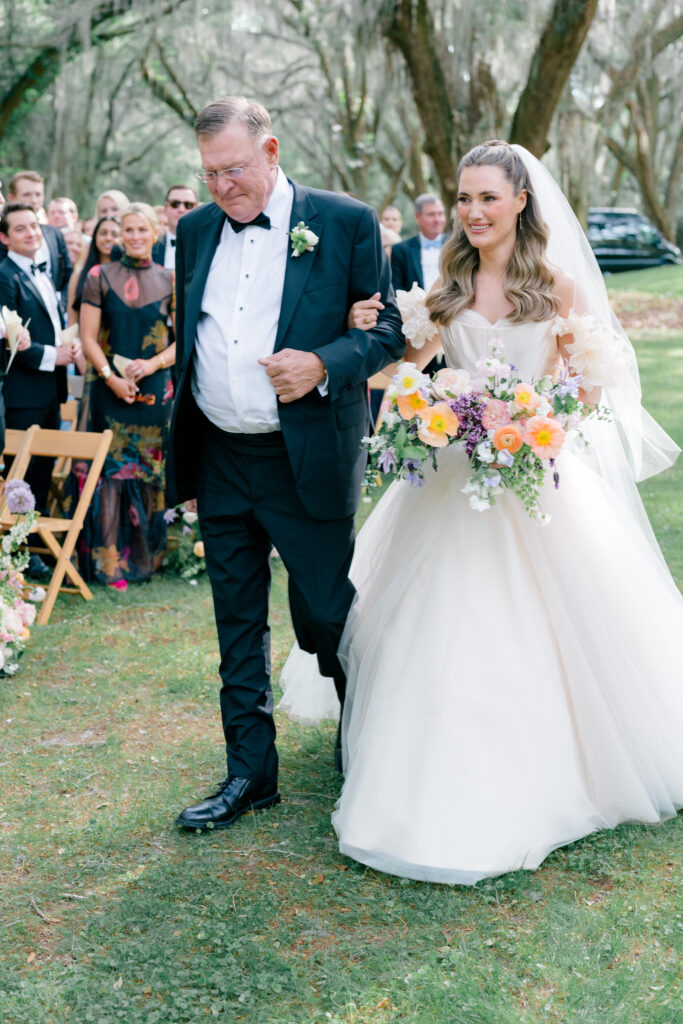 Bride in blush pink dress and purple and orange flowers escorted up the aisle by her dad. 