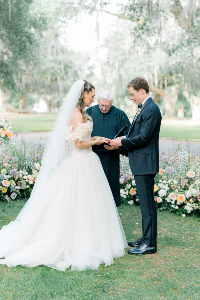 Wedding ring exchange. Groom places ring on brides finger. Wedding dress with decorative floral sleeves. 