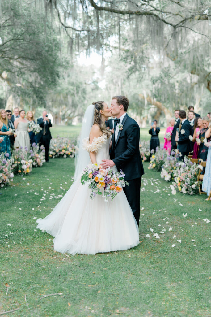 Bride and groom stop and kiss at outdoor wedding ceremony under spanish moss on Spring Island. 