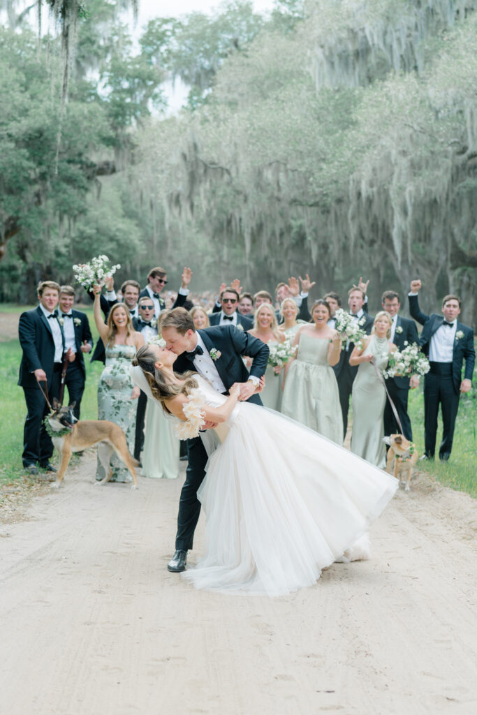 Groom dips bride and kisses her on dirt road with wedding party celebrating in the background. Spring Island destination wedding. 