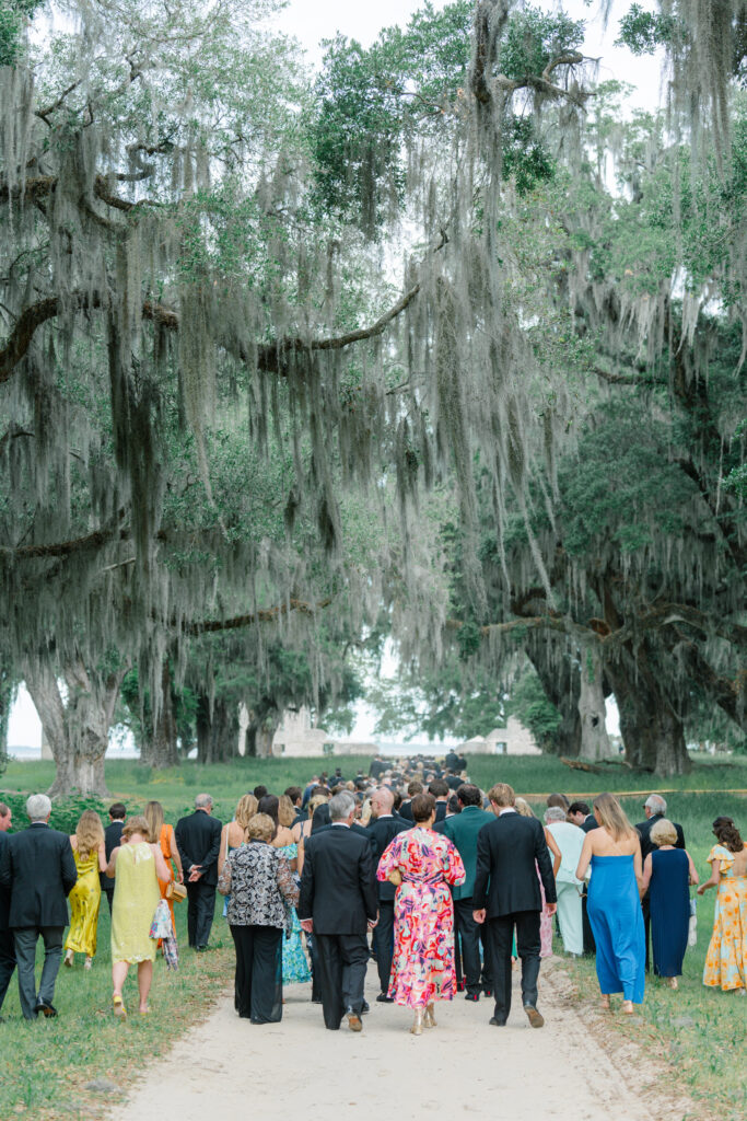 Wedding guests walking to cocktail hour on dirt road with spanish moss and live oak trees. 