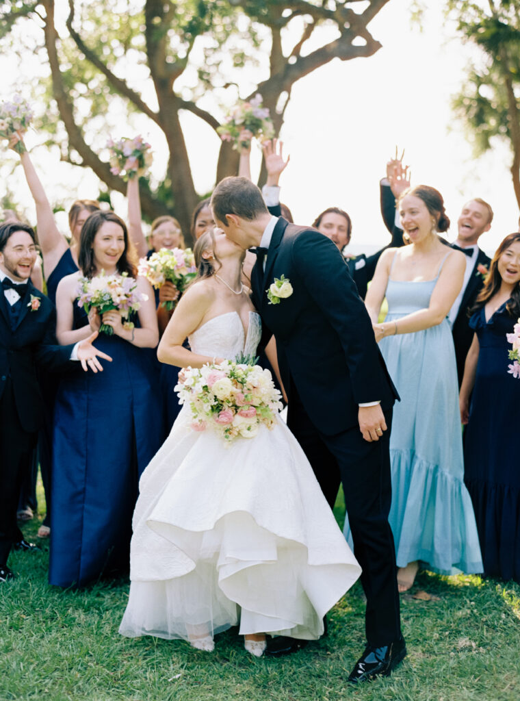 Bride and groom kiss with bridal party in the background. 