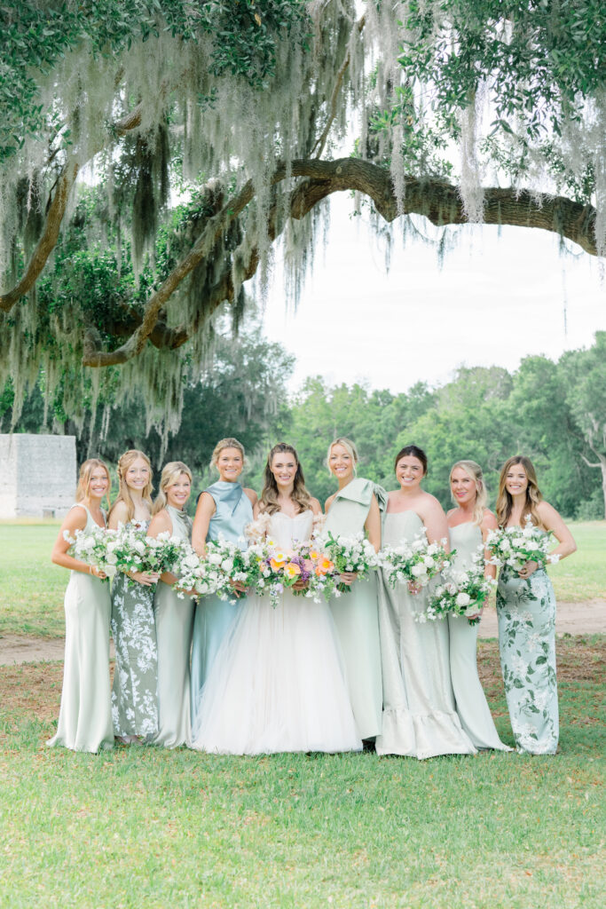 Bride and bridesmaids standing under spanish moss. Bridesmaids in mix-matched green dresses with flower patterns. 