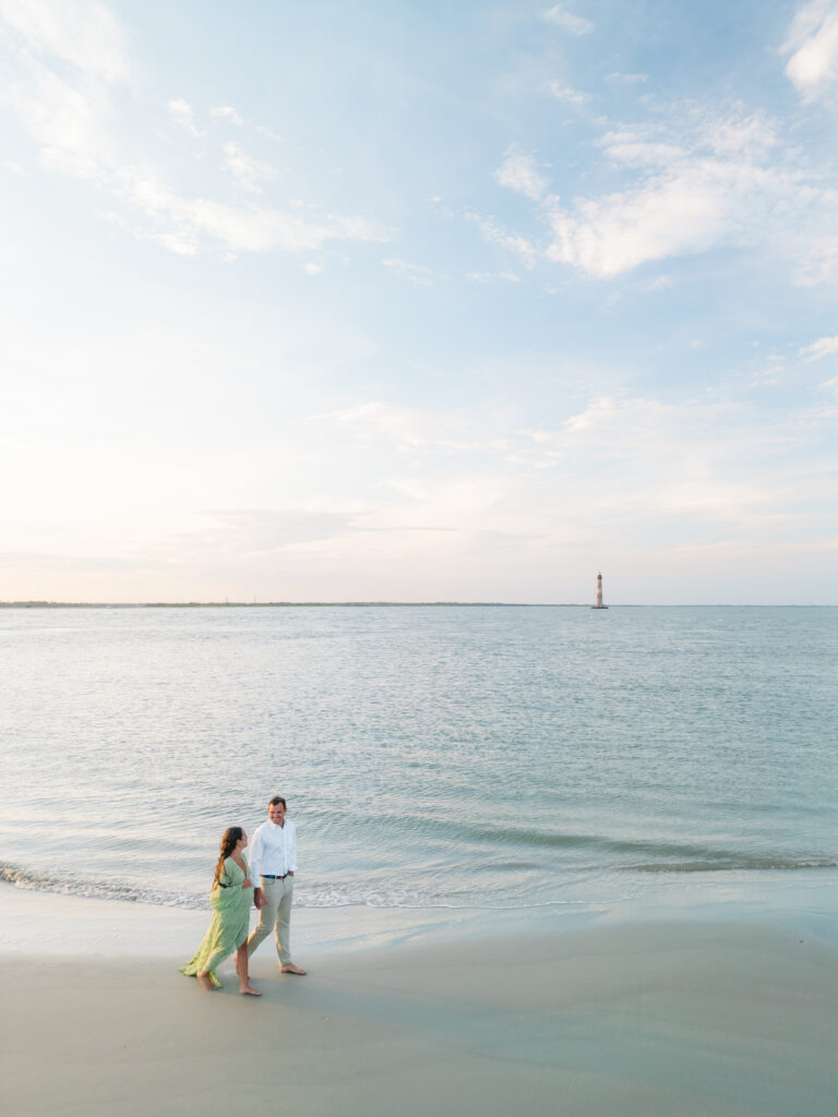 Couple holding hands on the beach with Morris Island Lighthouse in background. 