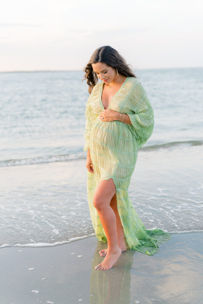 Maternity photos. Girl in sheer green dress holding and looking down at belly with the ocean water touching feet. Sunset maternity photos.