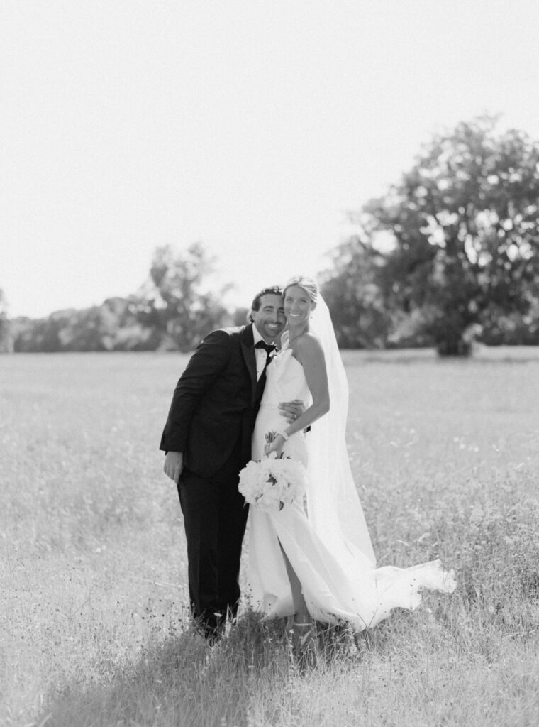 Black and white film photo of bride and groom standing in field with live oak trees in background. 