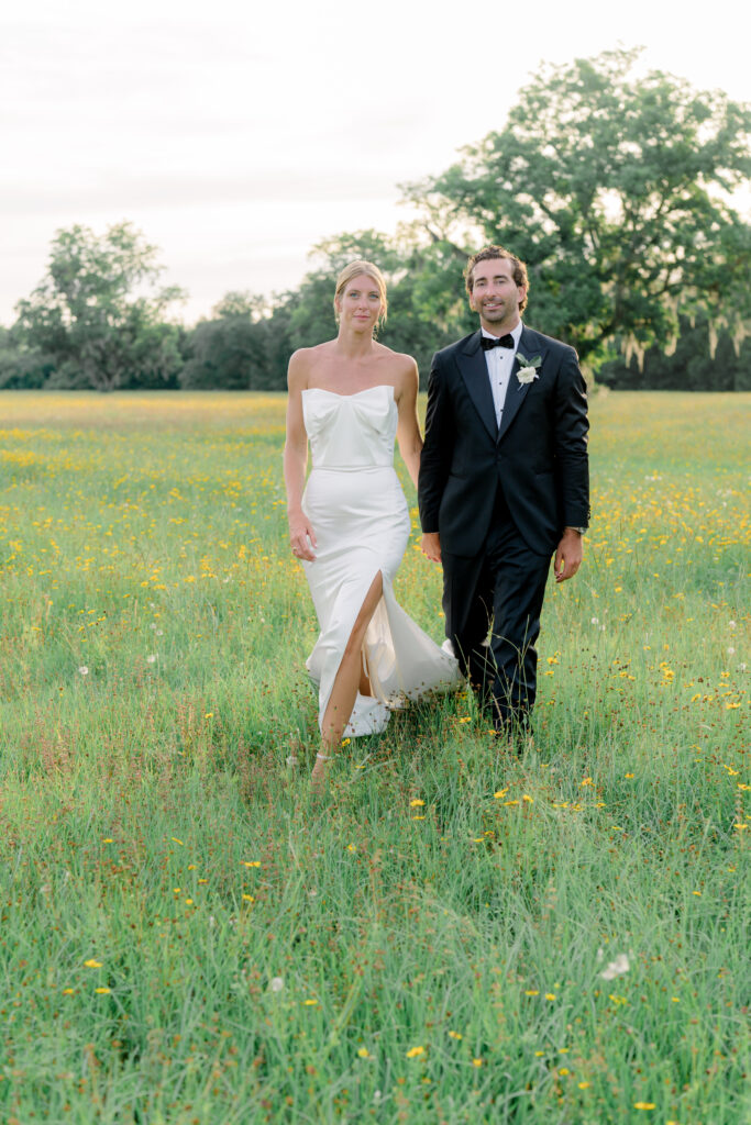 Bride and groom walking in field of yellow wildflowers. Beaufort waterfront wedding. 