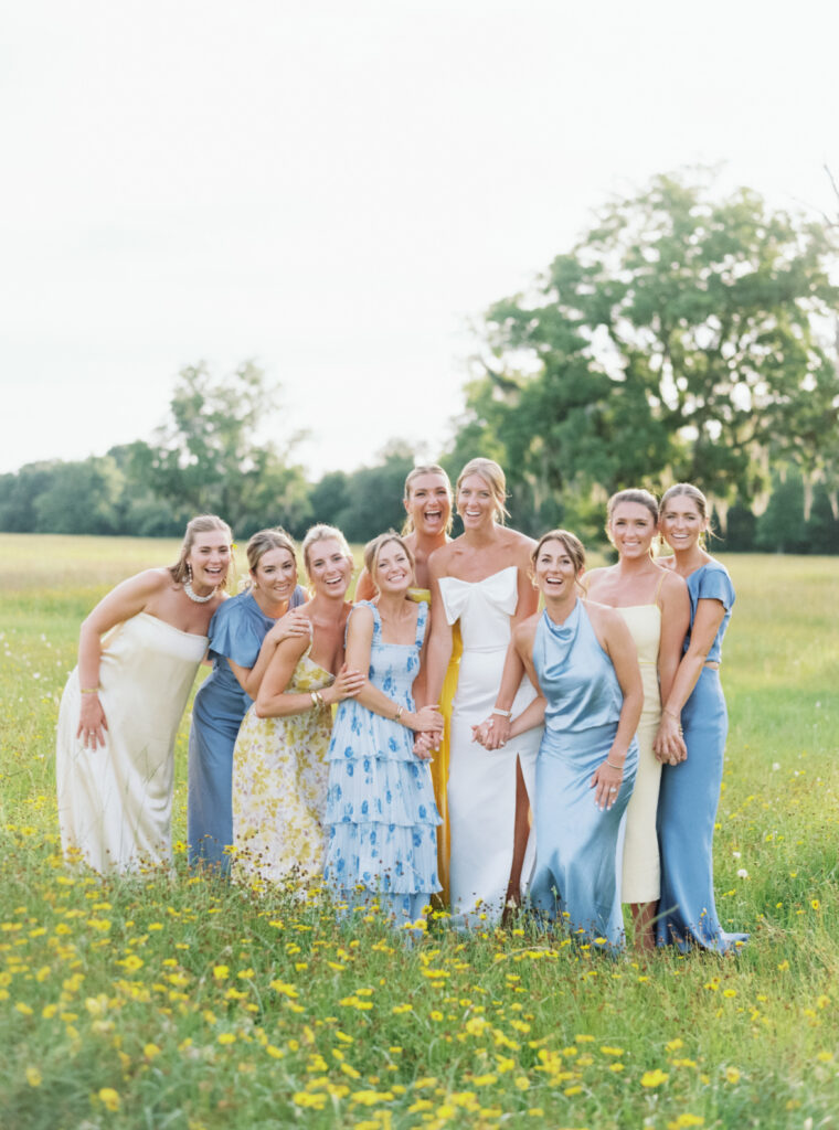 Bride and bridesmaids in the field at sunset with yellow wildflowers. Live oak tree with spanish moss in background. Blue and yellow dresses. 