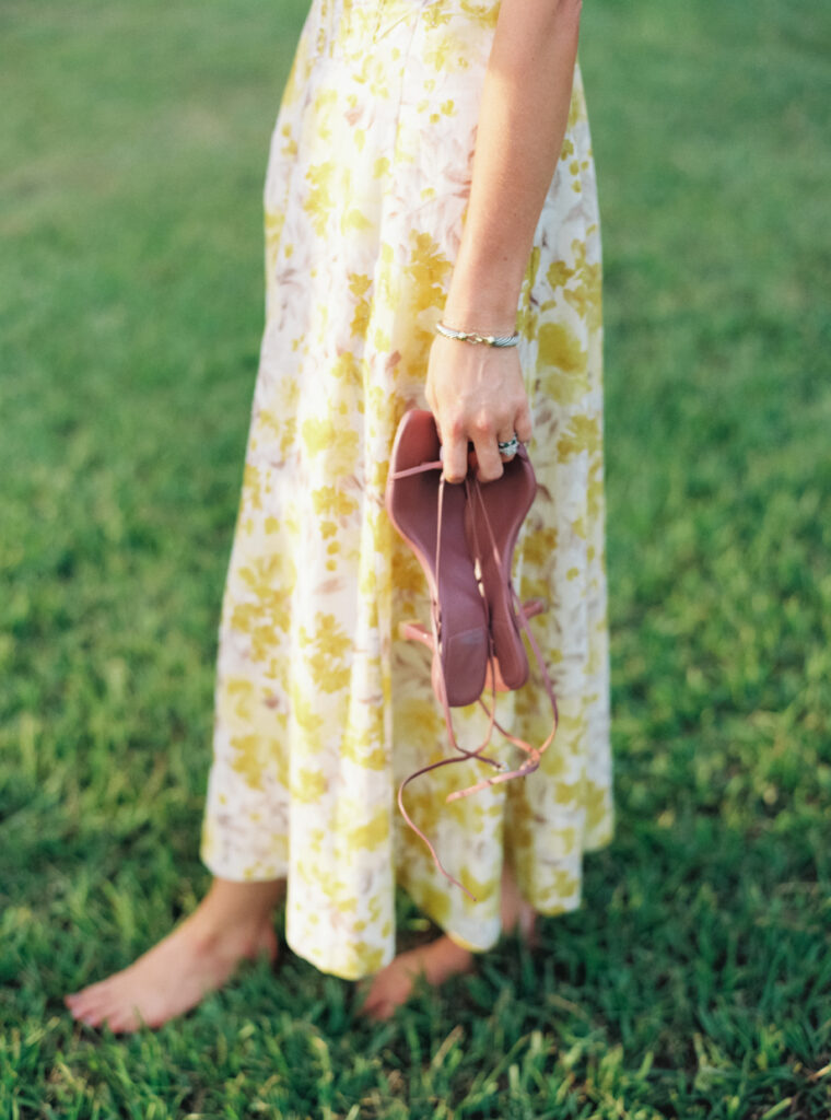 Bridesmaid in yellow dress holding strap heels in green grass. Wedding film photographer. 