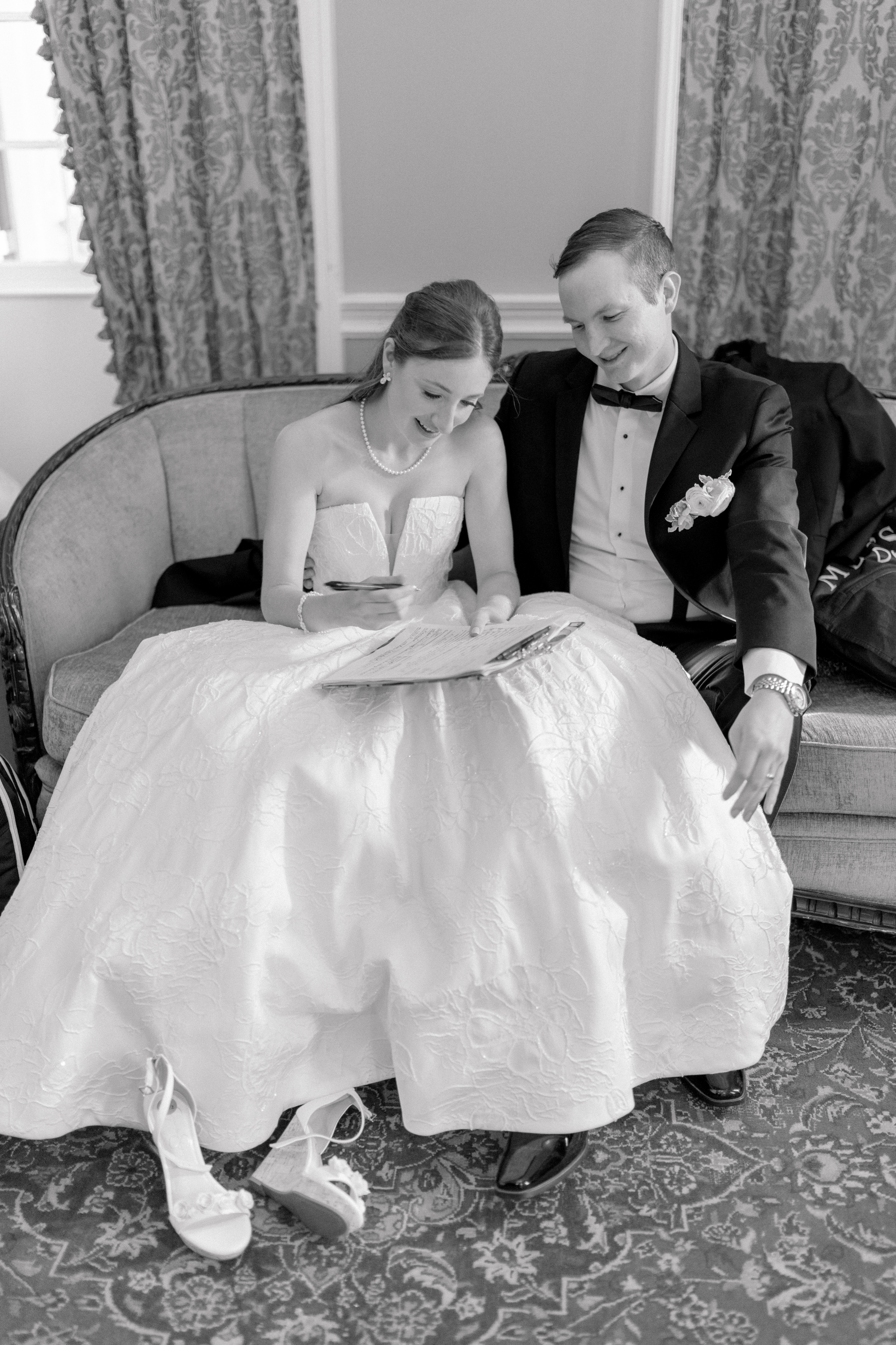 Black and white photo of bride and groom sitting on the couch and signing marriage license. Bride kicks off her heels. 