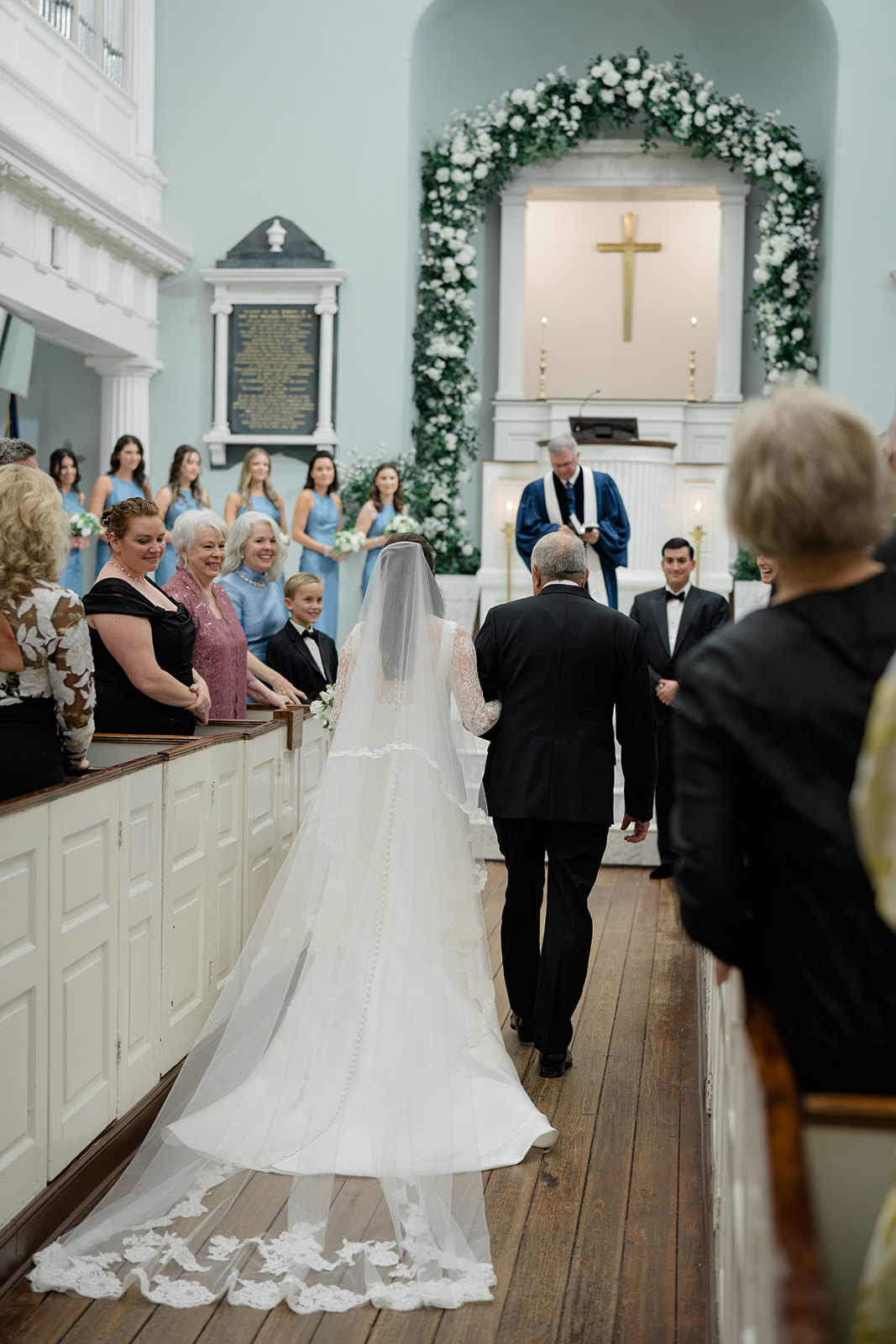 Wedding guests smile at bride and her dad as they enter wedding ceremony with big floral arch in background.