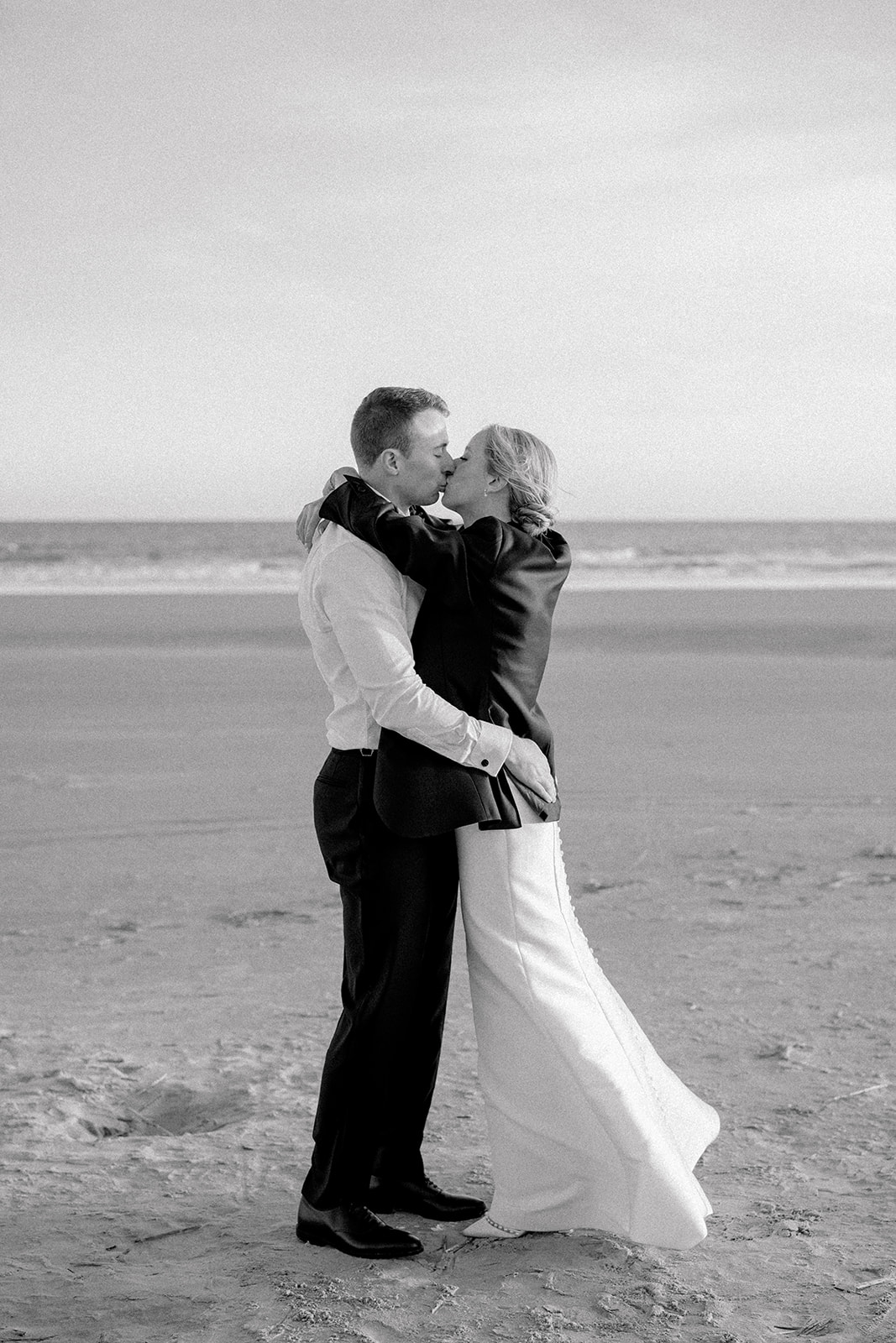 Black and white photo of bride and groom kissing with bride wearing grooms tuxedo jacket. 