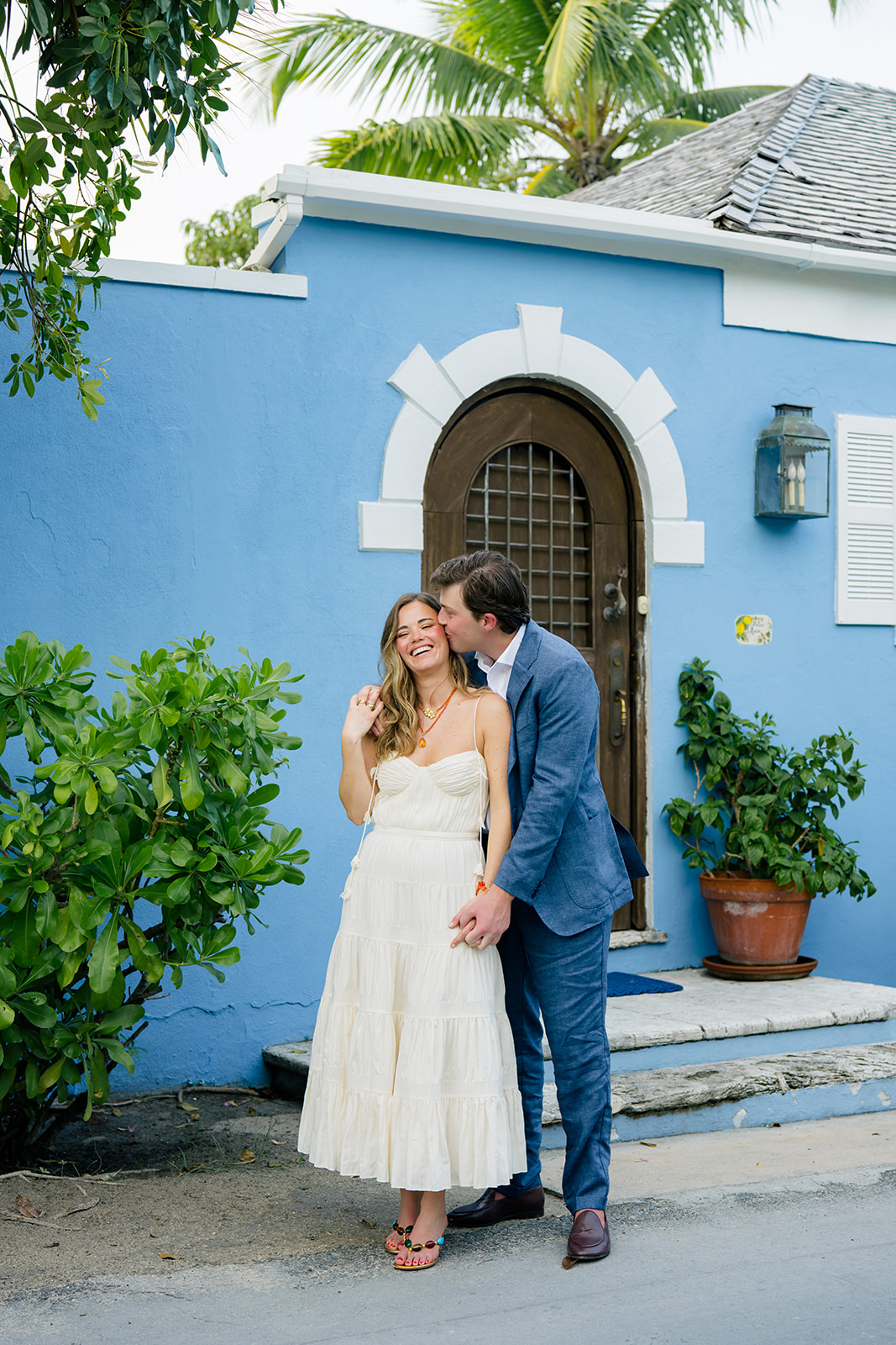 Groom leans in to kiss the cheek of the bride. Standing in front of light blue house with arched door and palm tree in background. 
