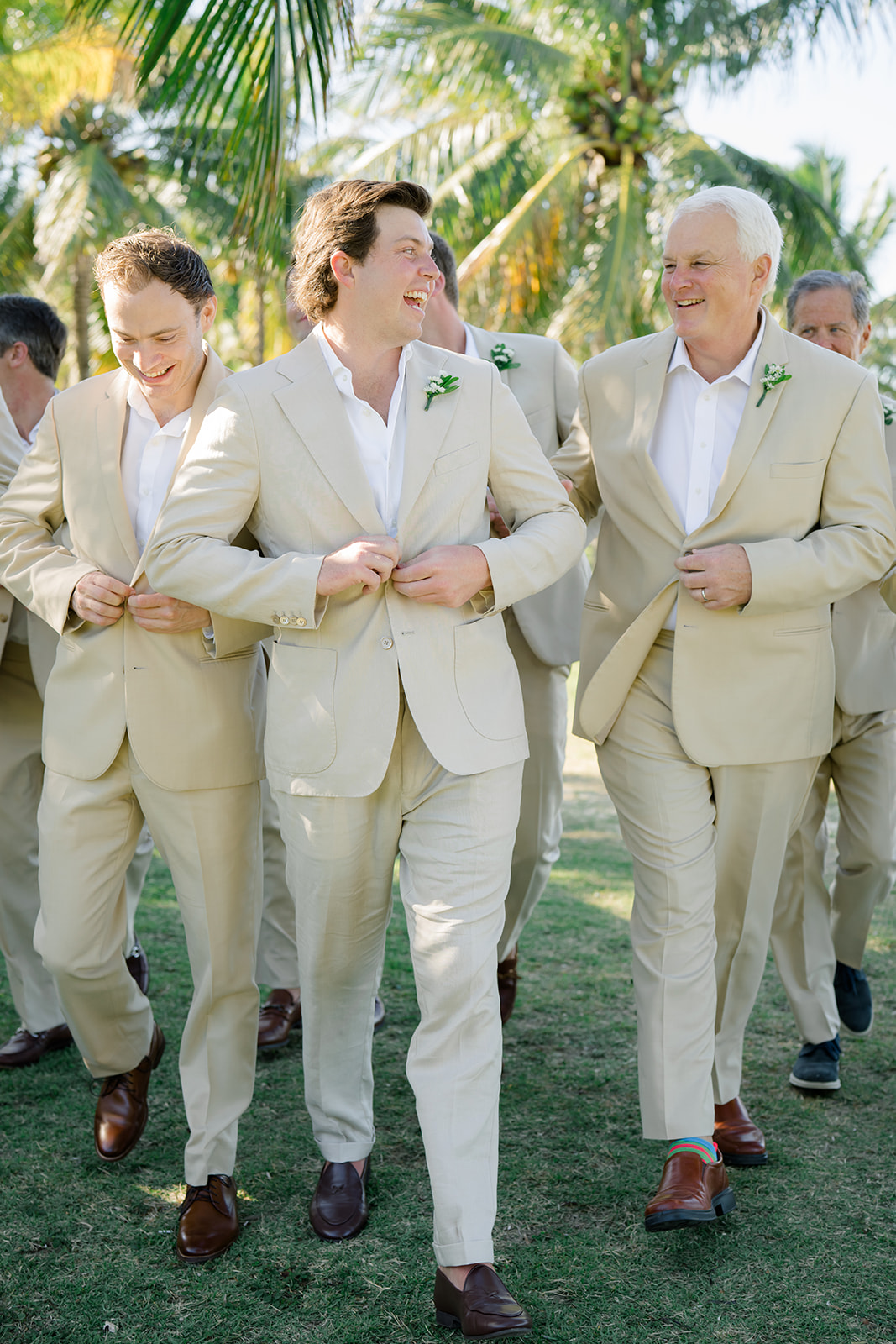 Groom walking with groomsmen, all wearing tan suits with white shirts and no collar. 