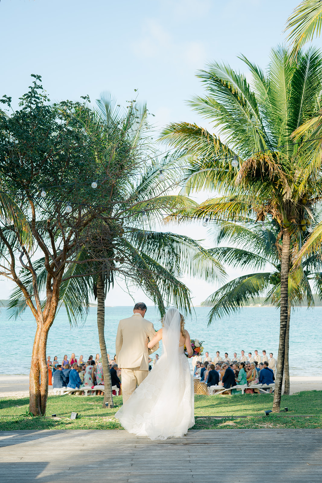 Father of the bride escorts daughter to beach front wedding ceremony. Palm trees in background with blue water. 