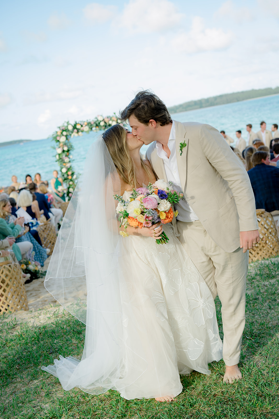 Bride and groom kiss after wedding with ceremony in background. 