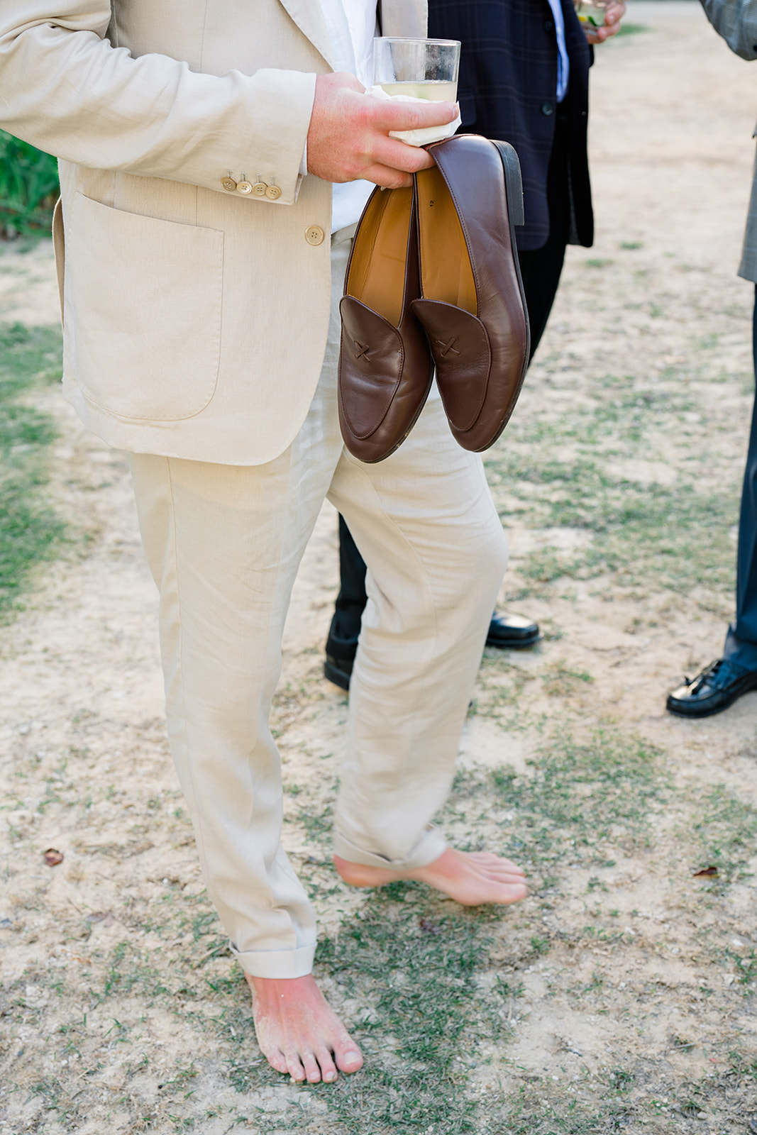Groom holds his loafers and a drink during cocktail hour. 