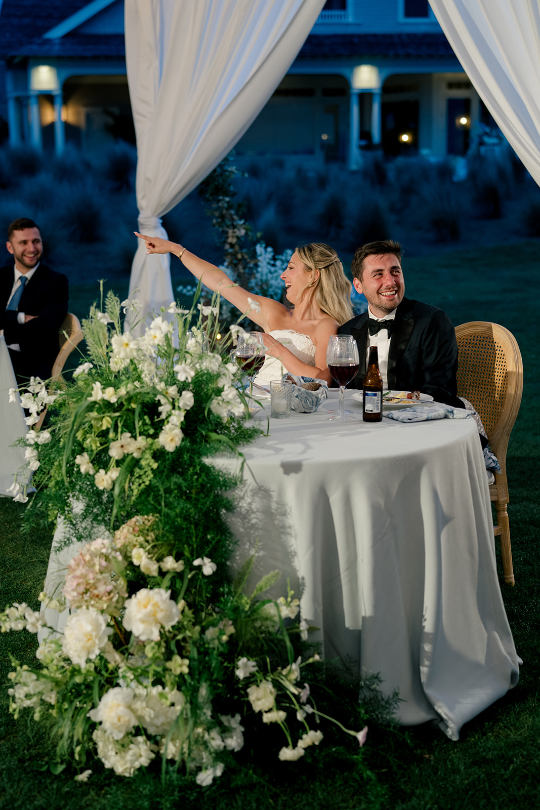 Bride laughs and points to a friend during speech at wedding reception. 
