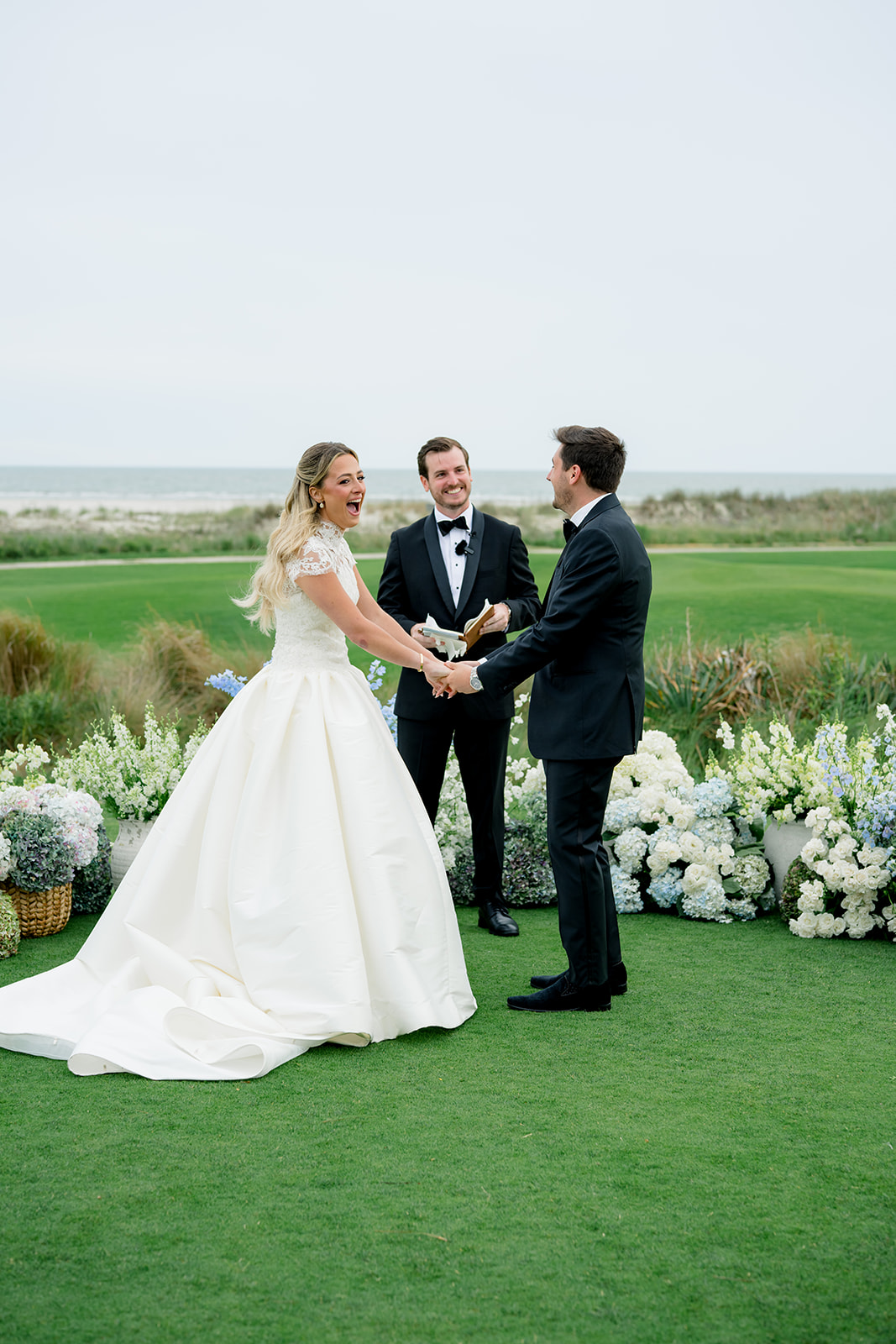Bride and groom laugh with the officiant. Blue and white flowers.